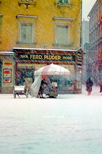 Heisse Maronen or Roasted Chestnuts seller in a snow storm in front of the Ferdinand Pluder Fashion Mode Shop. Austria 1939