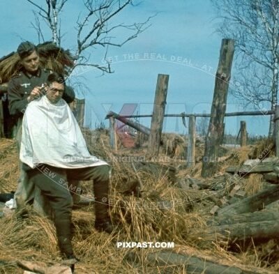 German army soldier getting his hair cut from field hard dressers. Beside destroyed farm house. Russia 1942
