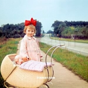 Little German girl with red hair ribbon with baby pram and dolly beside road into Siedlung Dessau Torten Germany 1939