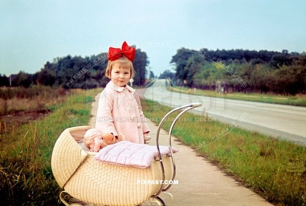 Little German girl with red hair ribbon with baby pram and dolly beside road into Siedlung Dessau Torten Germany 1939